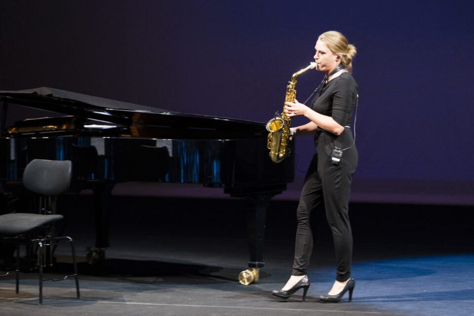 Eine Person steht auf einer Bühne und spielt Saxophon, gekleidet in Schwarz und mit hochhackigen Schuhen. Im Hintergrund befinden sich ein Flügel und ein leerer Stuhl. Die Bühne ist dunkel gehalten, das Licht richtet sich auf die Musikerin.