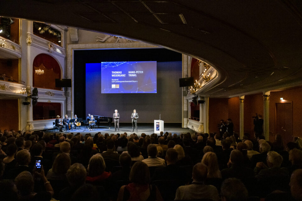 In einem prunkvoll dekorierten Theater mit Kronleuchtern und Balkonen findet eine formelle Veranstaltung statt. Auf der Bühne stehen zwei Personen vor einer großen Leinwand, während links eine kleine Musikgruppe spielt. Das Publikum sitzt und blickt zur Bühne.