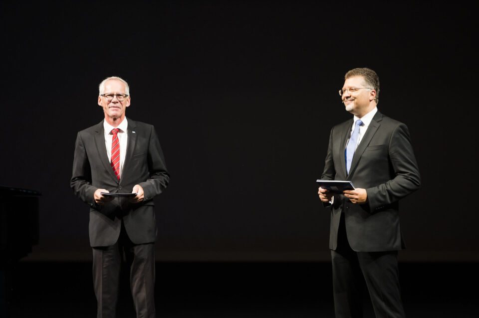 Zwei Personen stehen auf einer Bühne und tragen formelle Kleidung. Der Hintergrund ist dunkel.