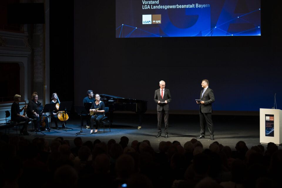 Auf einer Bühne stehen zwei Männer in Anzügen, einer mit Mikrofon, der andere mit einem Tablet. Links daneben sitzt ein Ensemble mit verschiedenen Musikinstrumenten wie Klavier, Violine, Cello und Klarinette, im Hintergrund ist eine große Leinwand mit Logos und Text zu sehen, davor das Publikum.
