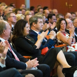 In einem Konferenzsaal oder Auditorium sitzt eine große Gruppe von Menschen in formeller Kleidung. Viele der Anwesenden applaudieren.