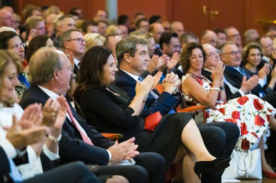 In einem Konferenzsaal oder Auditorium sitzt eine große Gruppe von Menschen in formeller Kleidung. Viele der Anwesenden applaudieren.