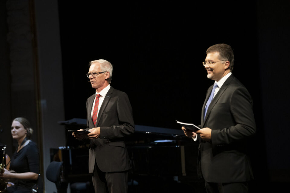 Zwei Personen stehen auf einer Bühne und tragen formelle Kleidung, beide halten Papiere in der Hand. Im Hintergrund ist eine weitere Person teilweise sichtbar, möglicherweise mit einem Musikinstrument. Die dunkle Umgebung deutet auf eine Veranstaltung oder Aufführung in einem Innenraum hin.