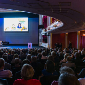Ein großes Publikum sitzt in einem prunkvoll dekorierten Theater mit roten Vorhängen und goldenen Verzierungen und blickt auf eine Bühne. Auf der Bühne befindet sich eine Leinwand, daneben spielt ein Ensemble mit Klavier und Streichinstrumenten.