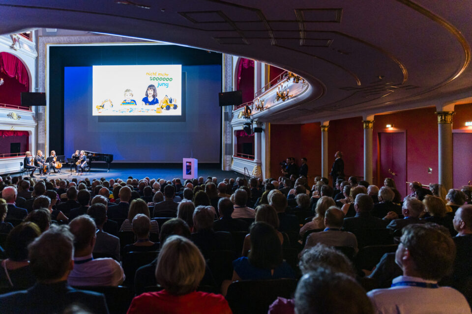 Ein großes Publikum sitzt in einem prunkvoll dekorierten Theater mit roten Vorhängen und goldenen Verzierungen und blickt auf eine Bühne. Auf der Bühne befindet sich eine Leinwand, daneben spielt ein Ensemble mit Klavier und Streichinstrumenten.