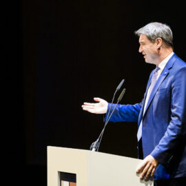 Eine Person steht hinter einem Rednerpult und hält eine Rede oder Präsentation. Sie trägt einen blauen Anzug mit Krawatte, gestikuliert mit einer Hand und hält sich mit der anderen am Pult fest. Zwei Mikrofone sind am Pult befestigt, der Hintergrund ist dunkel.