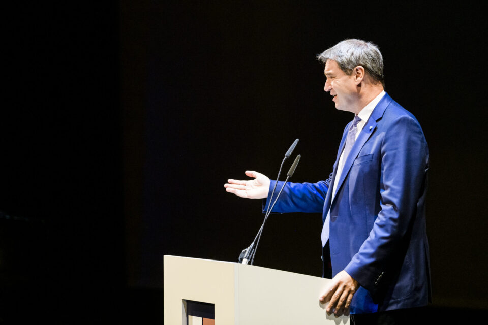 Eine Person steht hinter einem Rednerpult und hält eine Rede oder Präsentation. Sie trägt einen blauen Anzug mit Krawatte, gestikuliert mit einer Hand und hält sich mit der anderen am Pult fest. Zwei Mikrofone sind am Pult befestigt, der Hintergrund ist dunkel.