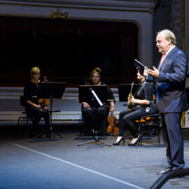 Auf einer Bühne sitzen vier Musiker mit Instrumenten wie Violine und Saxophon. Rechts steht ein Mann im Anzug mit Papieren oder einem Buch in der Hand. Der Hintergrund zeigt ein prunkvolles Theater mit roten Vorhängen und goldenen Verzierungen.