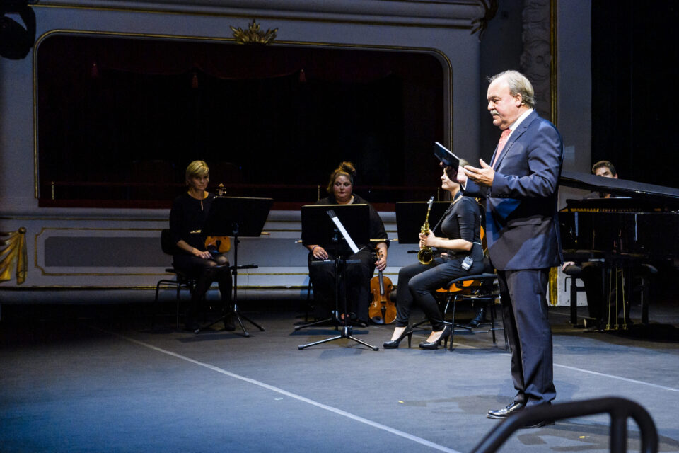 Auf einer Bühne sitzen vier Musiker mit Instrumenten wie Violine und Saxophon. Rechts steht ein Mann im Anzug mit Papieren oder einem Buch in der Hand. Der Hintergrund zeigt ein prunkvolles Theater mit roten Vorhängen und goldenen Verzierungen.