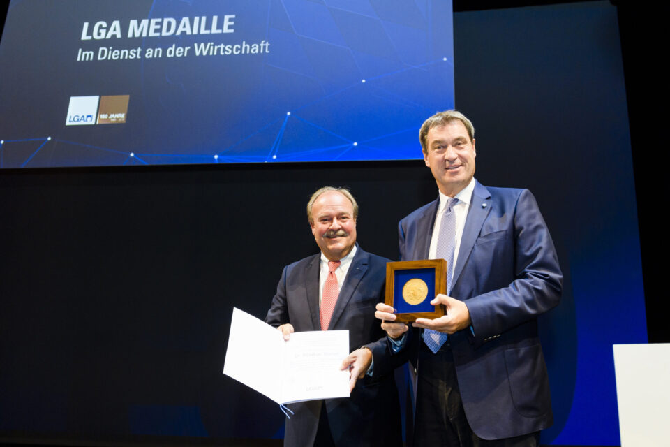 Zwei Personen stehen auf einer Bühne, eine hält eine geöffnete Urkunde oder ein Dokument, die andere eine gerahmte Medaille. Im Hintergrund ist eine große Leinwand mit einem Schriftzug und Logos zu sehen.