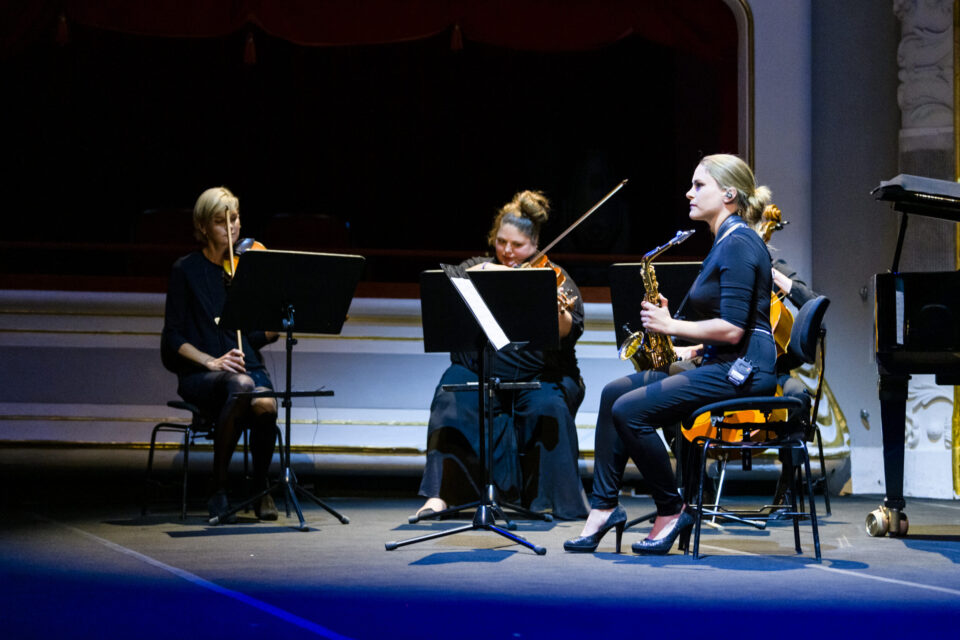 Drei Musiker sitzen auf einer Bühne und spielen verschiedene Instrumente: links ein Streichinstrument, in der Mitte ein Cello und rechts ein Saxophon. Vor ihnen stehen Notenständer, die Bühne ist in dunklem Licht mit blauen Akzenten beleuchtet.