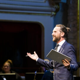 Eine Person in einem formellen Anzug steht mit einem Tablet oder Notizbuch auf einer Bühne oder vor einem Publikum. Im Hintergrund ist ein Teil eines prunkvollen Theaters mit dekorativen Elementen sowie eine weitere Person teilweise sichtbar.