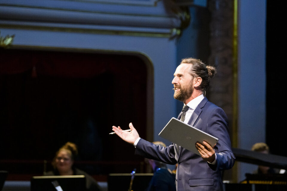 Eine Person in einem formellen Anzug steht mit einem Tablet oder Notizbuch auf einer Bühne oder vor einem Publikum. Im Hintergrund ist ein Teil eines prunkvollen Theaters mit dekorativen Elementen sowie eine weitere Person teilweise sichtbar.