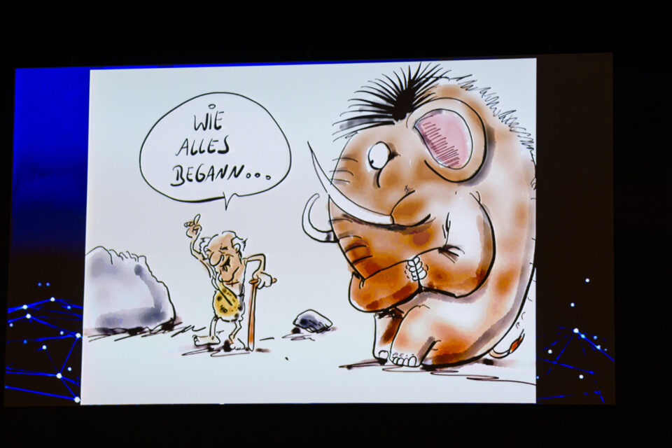 Das Bild zeigt eine Cartoon-Zeichnung mit einem älteren Höhlenmenschen, der sich mit einem Stock an einen großen wolligen Mammut wendet. Im Hintergrund sind Felsen zu sehen, die Szene spielt in einer prähistorischen Umgebung.