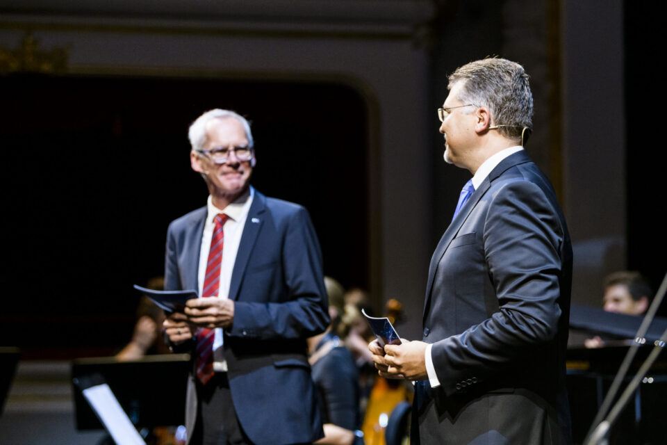 Zwei Personen in Anzügen stehen auf einer Bühne, beide halten Papiere in der Hand. Im Hintergrund sind Musikinstrumente und weitere Personen zu sehen, was auf eine Festveranstaltung hinweist.