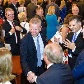 Eine große Gruppe von Menschen in formeller Kleidung ist bei einer gesellschaftlichen oder beruflichen Veranstaltung versammelt. Viele Personen unterhalten sich, geben sich die Hand oder interagieren miteinander.