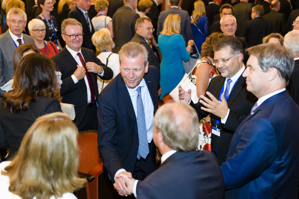 Eine große Gruppe von Menschen in formeller Kleidung ist bei einer gesellschaftlichen oder beruflichen Veranstaltung versammelt. Viele Personen unterhalten sich, geben sich die Hand oder interagieren miteinander.
