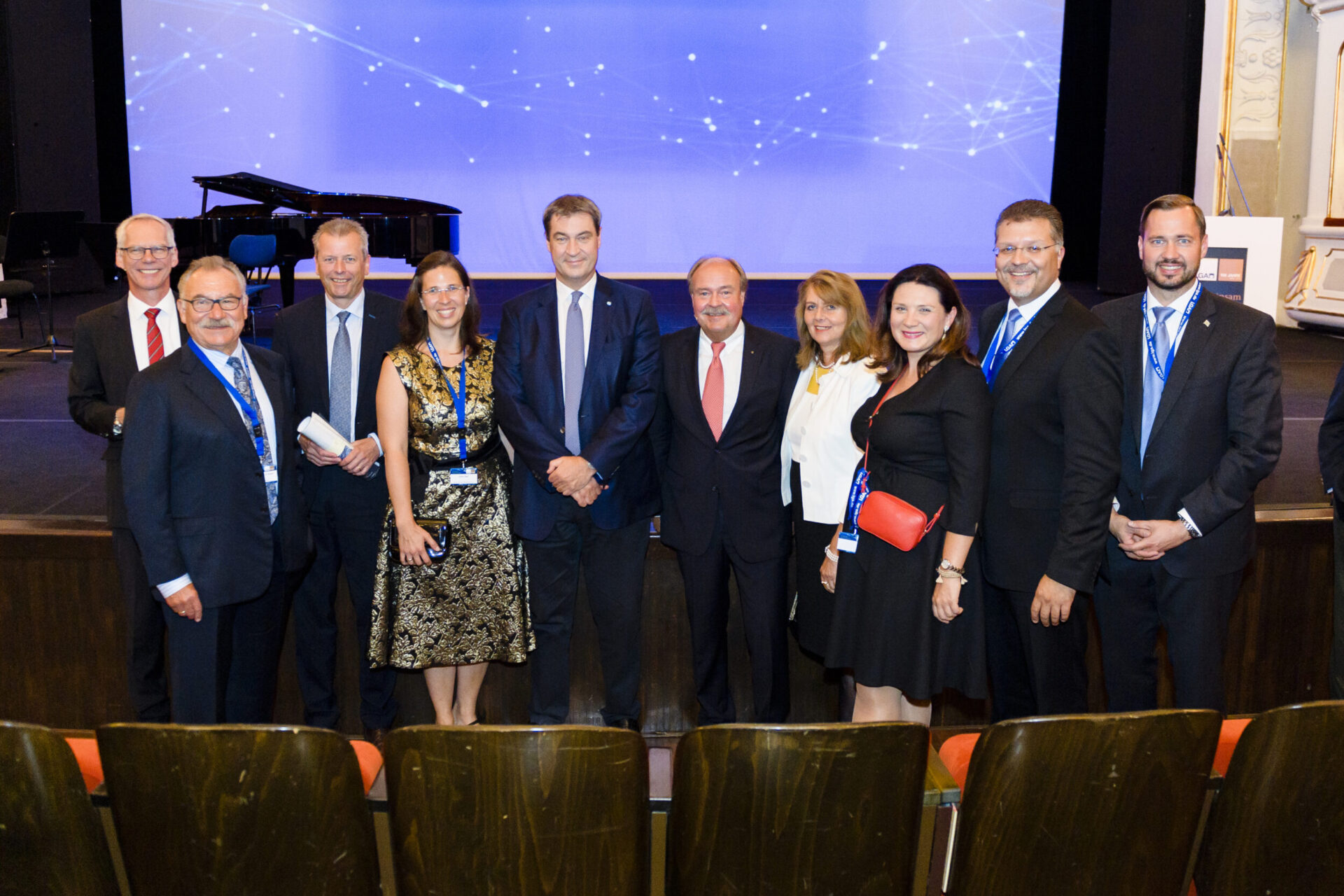 Zehn Personen auf einer Bühne, alle tragen formelle Kleidung wie Anzüge, Krawatten oder Businesskleidung. Im Hintergrund ist eine blaue Leinwand mit weißen Lichtmustern zu sehen, links steht ein Flügel. Alle Personen tragen Namensschilder an einem Band um den Hals, was auf eine Veranstaltung hinweist.
