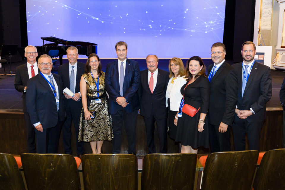 Zehn Personen auf einer Bühne, alle tragen formelle Kleidung wie Anzüge, Krawatten oder Businesskleidung. Im Hintergrund ist eine blaue Leinwand mit weißen Lichtmustern zu sehen, links steht ein Flügel. Alle Personen tragen Namensschilder an einem Band um den Hals, was auf eine Veranstaltung hinweist.