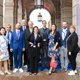 Zehn Personen stehen vor einem bogenförmigen Eingang mit steinernen Säulen, alle tragen formelle Kleidung wie Anzüge, Kleider oder Business-Outfits. Sie haben Namensschilder an einem Band um den Hals, was auf eine Veranstaltung oder Konferenz hinweist. Im Hintergrund hängt eine große Laterne von der Decke des Bogens.