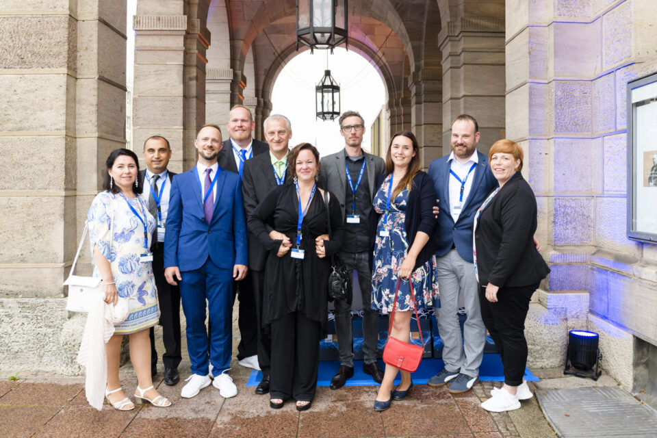 Zehn Personen stehen vor einem bogenförmigen Eingang mit steinernen Säulen, alle tragen formelle Kleidung wie Anzüge, Kleider oder Business-Outfits. Sie haben Namensschilder an einem Band um den Hals, was auf eine Veranstaltung oder Konferenz hinweist. Im Hintergrund hängt eine große Laterne von der Decke des Bogens.