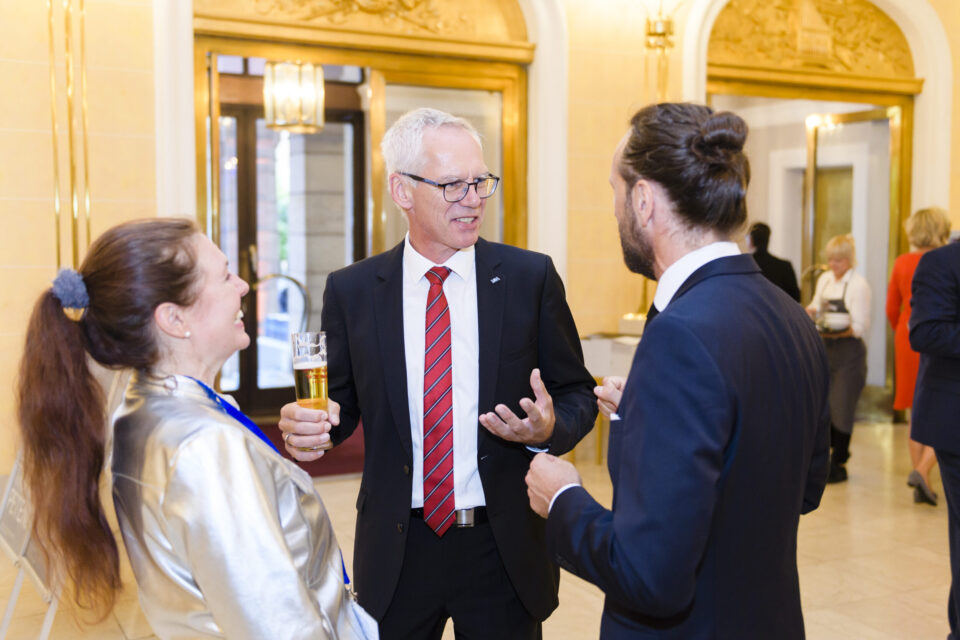 Drei Personen unterhalten sich bei einer formellen Veranstaltung, die mittlere Person hält ein Glas Bier und trägt einen schwarzen Anzug mit rot gestreifter Krawatte. Links steht eine Person in einer glänzenden silbernen Jacke, rechts eine Person im dunklen Anzug. Der Hintergrund zeigt eine prunkvolle Umgebung mit goldenen Verzierungen und Kronleuchtern, weitere Gäste sind im Raum verteilt.