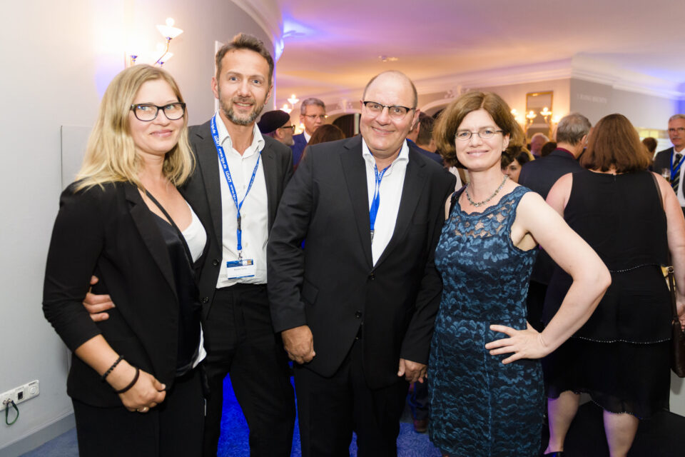 Eine Gruppe von Personen in formeller Kleidung befindet sich bei einer gesellschaftlichen oder beruflichen Veranstaltung in einem Innenraum. Im Hintergrund sind weitere Gäste und stimmungsvolle Beleuchtung zu sehen, was auf eine festliche oder formelle Atmosphäre hinweist.