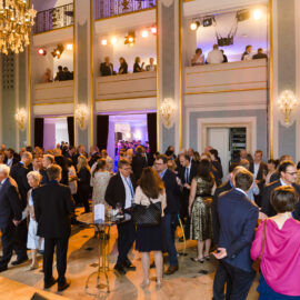 Das Bild zeigt eine große Menschenmenge bei einer Veranstaltung in einem eleganten Innenraum mit Kronleuchtern, Wandleuchten und zwei Etagen mit Balkonen, auf denen weitere Gäste stehen. Die Szene wirkt wie eine festliche Veranstaltung.