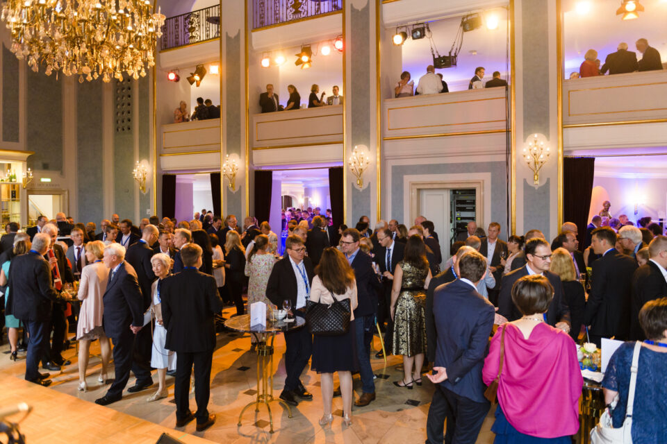 Das Bild zeigt eine große Menschenmenge bei einer Veranstaltung in einem eleganten Innenraum mit Kronleuchtern, Wandleuchten und zwei Etagen mit Balkonen, auf denen weitere Gäste stehen. Die Szene wirkt wie eine festliche Veranstaltung.