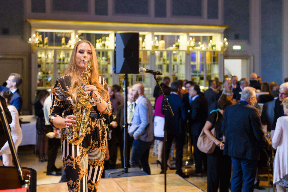 Das Bild zeigt eine gesellschaftliche Veranstaltung in einem großen, elegant dekorierten Raum mit warmem Licht. Im Vordergrund spielt eine Person Saxophon, im Hintergrund unterhalten sich mehrere Gäste an Tischen mit Getränken und Speisen. Ein Mikrofonständer und ein Lautsprecher sind ebenfalls sichtbar.