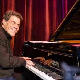 Eine Person in einem schwarzen Anzug spielt auf einem Flügel, das Gesicht ist verpixelt. Im Hintergrund hängen rote Vorhänge, was auf eine Aufführung oder Veranstaltung auf einer Bühne hinweist.