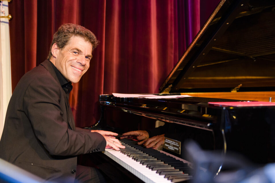 Eine Person in einem schwarzen Anzug spielt auf einem Flügel, das Gesicht ist verpixelt. Im Hintergrund hängen rote Vorhänge, was auf eine Aufführung oder Veranstaltung auf einer Bühne hinweist.