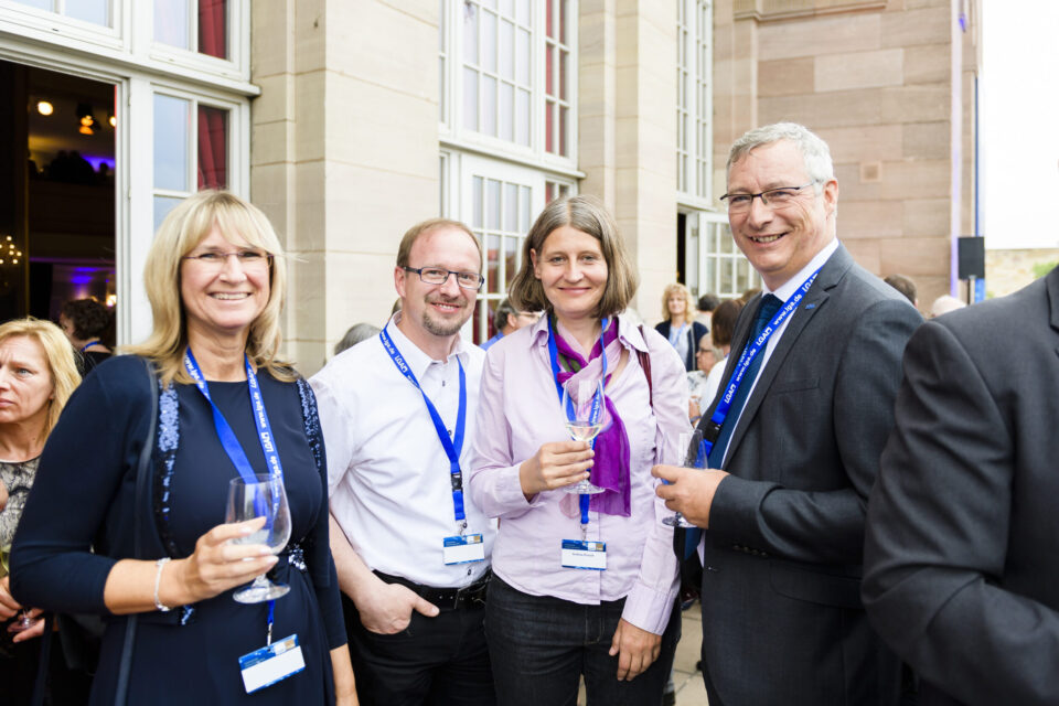 Fünf Personen stehen bei einer formellen oder beruflichen Veranstaltung zusammen, alle tragen Namensschilder an blauen Bändern um den Hals. Drei von ihnen halten Weingläser in der Hand. Im Hintergrund sind große Fenster und ein Gebäude aus Stein zu sehen.