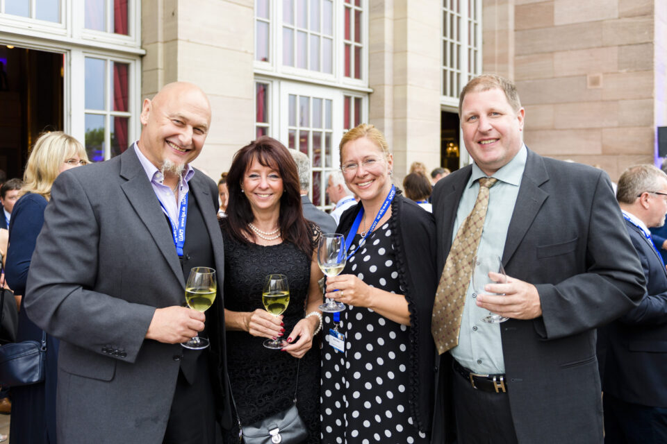Vier Personen stehen bei einer gesellschaftlichen oder formellen Veranstaltung zusammen, alle tragen elegante Kleidung und halten ein Glas Weißwein in der Hand. Im Hintergrund sind große Fenster und Steinwände zu sehen, was auf einen stilvollen Veranstaltungsort hinweist. Weitere Gäste unterhalten sich im Hintergrund.