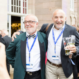 Zwei Personen stehen eng beieinander, eine legt den Arm um die Schulter der anderen. Beide tragen formelle Kleidung mit blauen Namensbändern, eine Person hält zwei Gläser Bier. Im Hintergrund sind weitere Gäste in festlicher Kleidung, ein Gebäude mit großen Fenstern und eine Person, die mit einem Smartphone ein Foto macht.