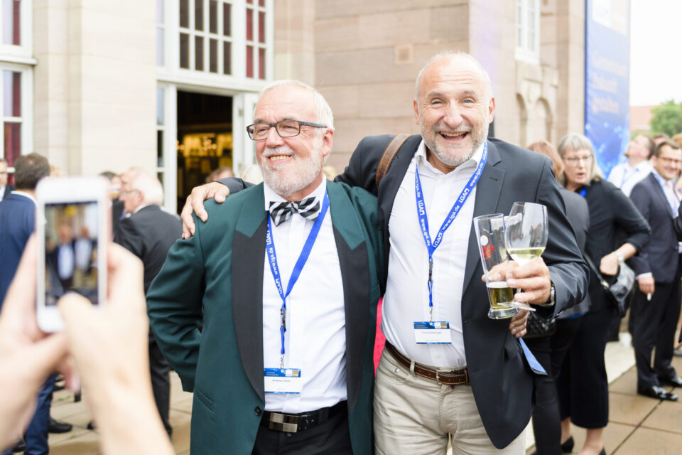 Zwei Personen stehen eng beieinander, eine legt den Arm um die Schulter der anderen. Beide tragen formelle Kleidung mit blauen Namensbändern, eine Person hält zwei Gläser Bier. Im Hintergrund sind weitere Gäste in festlicher Kleidung, ein Gebäude mit großen Fenstern und eine Person, die mit einem Smartphone ein Foto macht.