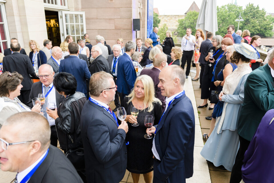 Eine große Gruppe von Personen hat sich bei einer Veranstaltung im Freien versammelt, viele tragen formelle Kleidung und halten Weingläser in der Hand. Die meisten haben Namensschilder an blauen Bändern um den Hals. Der Veranstaltungsort scheint eine Terrasse oder ein Hof neben einem Gebäude mit großen Fenstern und Steinwänden zu sein.