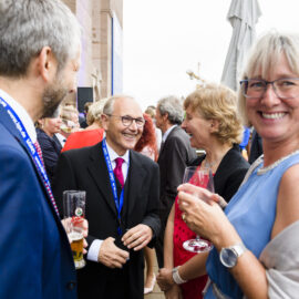 Eine Gruppe von Personen befindet sich bei einer Veranstaltung im Freien. Sie tragen Anzüge, Kleider und andere festliche Kleidung, einige halten Getränke wie Bier oder Wein in der Hand. Im Hintergrund sind Gebäude und ein Sonnenschirm zu sehen.