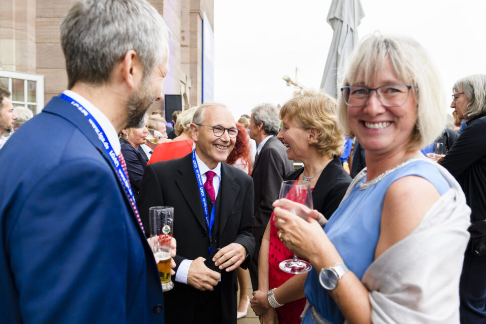 Eine Gruppe von Personen befindet sich bei einer Veranstaltung im Freien. Sie tragen Anzüge, Kleider und andere festliche Kleidung, einige halten Getränke wie Bier oder Wein in der Hand. Im Hintergrund sind Gebäude und ein Sonnenschirm zu sehen.