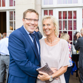 Im Vordergrund stehen ein Mann im blauen Anzug mit Krawatte und eine Frau im hellen Kleid, er hat den Arm um sie gelegt. Im Hintergrund unterhalten sich weitere Personen, einige mit Getränken in der Hand, vor einem eleganten Gebäude mit großen Fenstern und Türen.