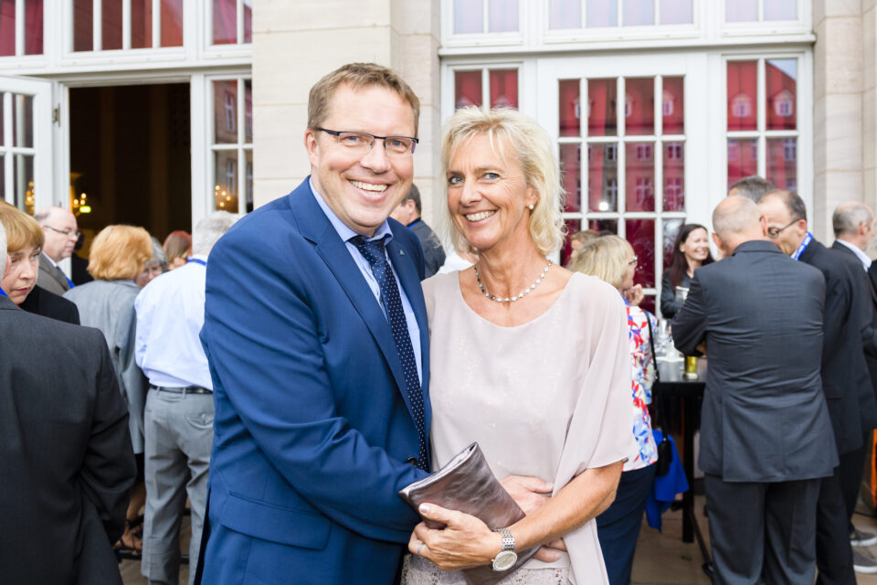 Im Vordergrund stehen ein Mann im blauen Anzug mit Krawatte und eine Frau im hellen Kleid, er hat den Arm um sie gelegt. Im Hintergrund unterhalten sich weitere Personen, einige mit Getränken in der Hand, vor einem eleganten Gebäude mit großen Fenstern und Türen.