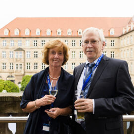Zwei Personen stehen auf einem Balkon und halten Getränke in der Hand, beide tragen formelle Kleidung – eine Person im Anzug mit Krawatte, die andere in einem dunklen Kleid. Sie tragen Namensschilder an Bändern um den Hals, was auf eine Veranstaltung oder Konferenz hinweist. Im Hintergrund ist ein großes Gebäude mit vielen Fenstern und rotem Ziegeldach zu sehen, das historisch oder institutionell wirkt.