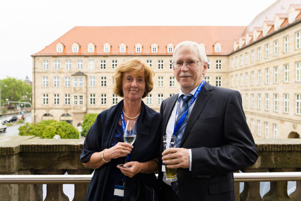 Zwei Personen stehen auf einem Balkon und halten Getränke in der Hand, beide tragen formelle Kleidung – eine Person im Anzug mit Krawatte, die andere in einem dunklen Kleid. Sie tragen Namensschilder an Bändern um den Hals, was auf eine Veranstaltung oder Konferenz hinweist. Im Hintergrund ist ein großes Gebäude mit vielen Fenstern und rotem Ziegeldach zu sehen, das historisch oder institutionell wirkt.