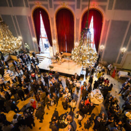 Das Bild zeigt einen großen, eleganten Saal mit hohen Decken, prunkvollen Kronleuchtern und großen roten Vorhängen vor hohen Fenstern. In der Mitte befindet sich eine Bühne, auf der Musiker auftreten. Die Gäste tragen formelle Kleidung und stehen in Gruppen an Tischen, unterhalten sich und genießen die Veranstaltung.