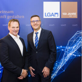 Zwei Personen in formeller Kleidung stehen vor einem Hintergrund mit grafischen Elementen aus Linien und Knoten. Auf dem Hintergrund sind Logos und Schriftzüge zu sehen.