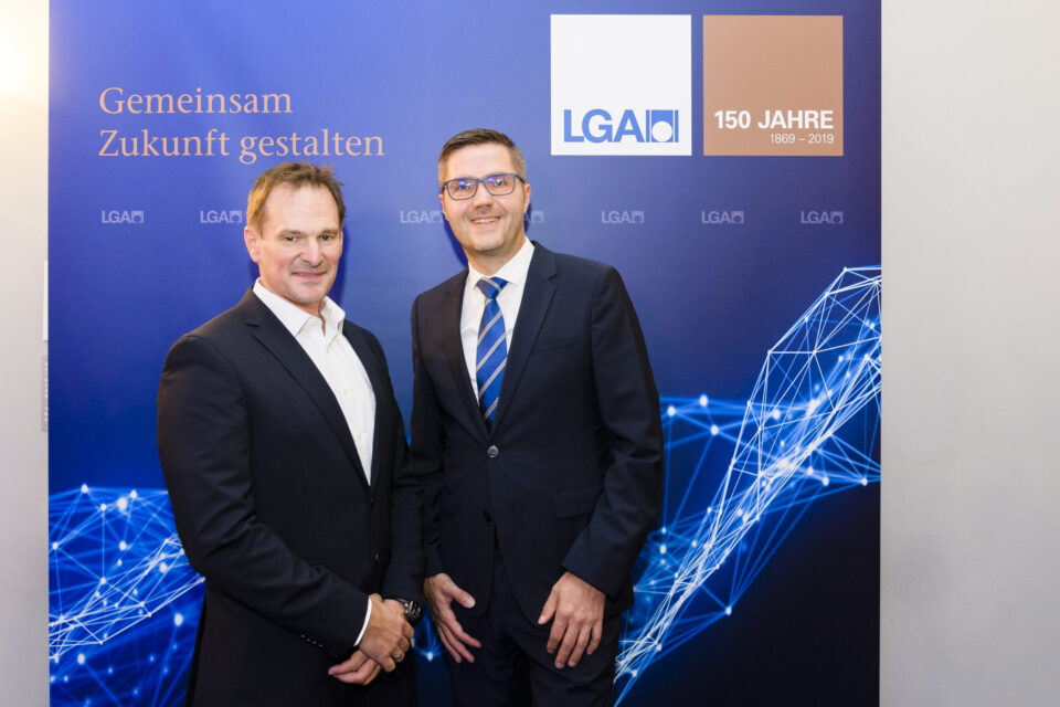 Zwei Personen in formeller Kleidung stehen vor einem Hintergrund mit grafischen Elementen aus Linien und Knoten. Auf dem Hintergrund sind Logos und Schriftzüge zu sehen.