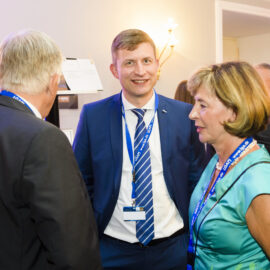 Drei Personen stehen im Vordergrund eines formellen Events oder einer Konferenz und unterhalten sich. Sie tragen Namensschilder mit blauen Bändern, was auf ihre Teilnahme an der Veranstaltung hinweist. Im Hintergrund sind weitere Personen und Wandbeleuchtung zu sehen.