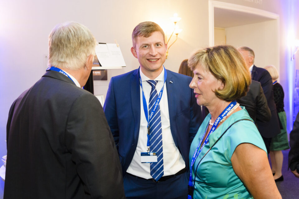 Drei Personen stehen im Vordergrund eines formellen Events oder einer Konferenz und unterhalten sich. Sie tragen Namensschilder mit blauen Bändern, was auf ihre Teilnahme an der Veranstaltung hinweist. Im Hintergrund sind weitere Personen und Wandbeleuchtung zu sehen.
