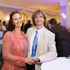 Ein elegant gekleidetes Paar steht Hand in Hand bei einer Veranstaltung und posiert für die Kamera. Die Frau trägt ein funkelndes, pfirsichfarbenes Kleid mit Perlenverzierungen, der Mann einen hellen Anzug mit gemusterter Krawatte. Im Hintergrund sind weitere Personen sowie ein Tisch mit Speisen zu sehen.