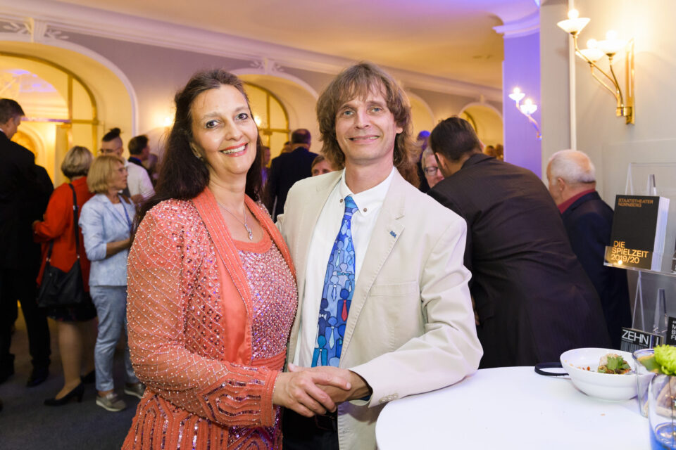 Ein elegant gekleidetes Paar steht Hand in Hand bei einer Veranstaltung und posiert für die Kamera. Die Frau trägt ein funkelndes, pfirsichfarbenes Kleid mit Perlenverzierungen, der Mann einen hellen Anzug mit gemusterter Krawatte. Im Hintergrund sind weitere Personen sowie ein Tisch mit Speisen zu sehen.